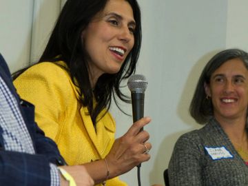 Woman in yellow jacket speaking into a microphone during a discussion panel